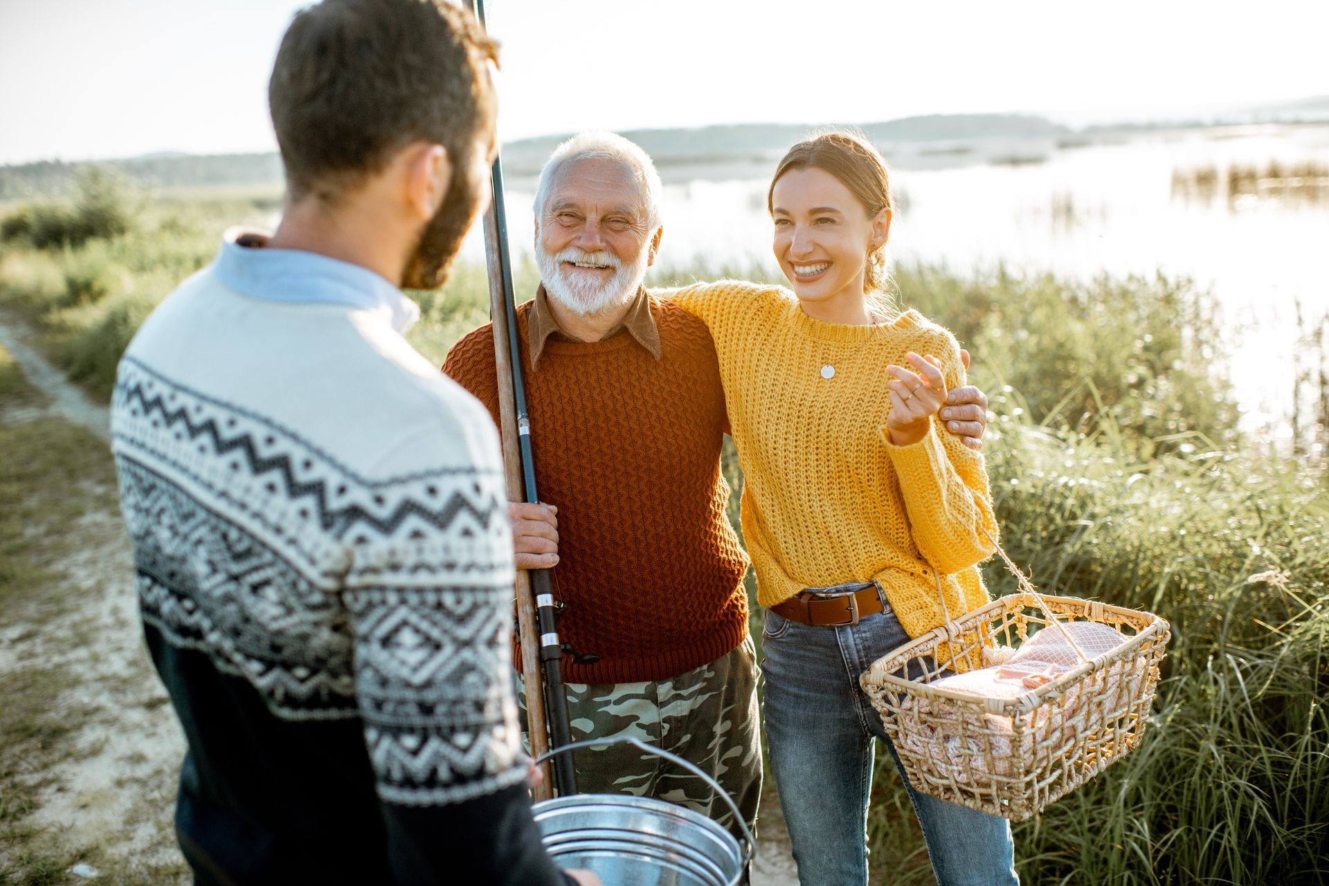 Three people stand by a lakeside, smiling and holding a fishing net and basket in bright sunlight.