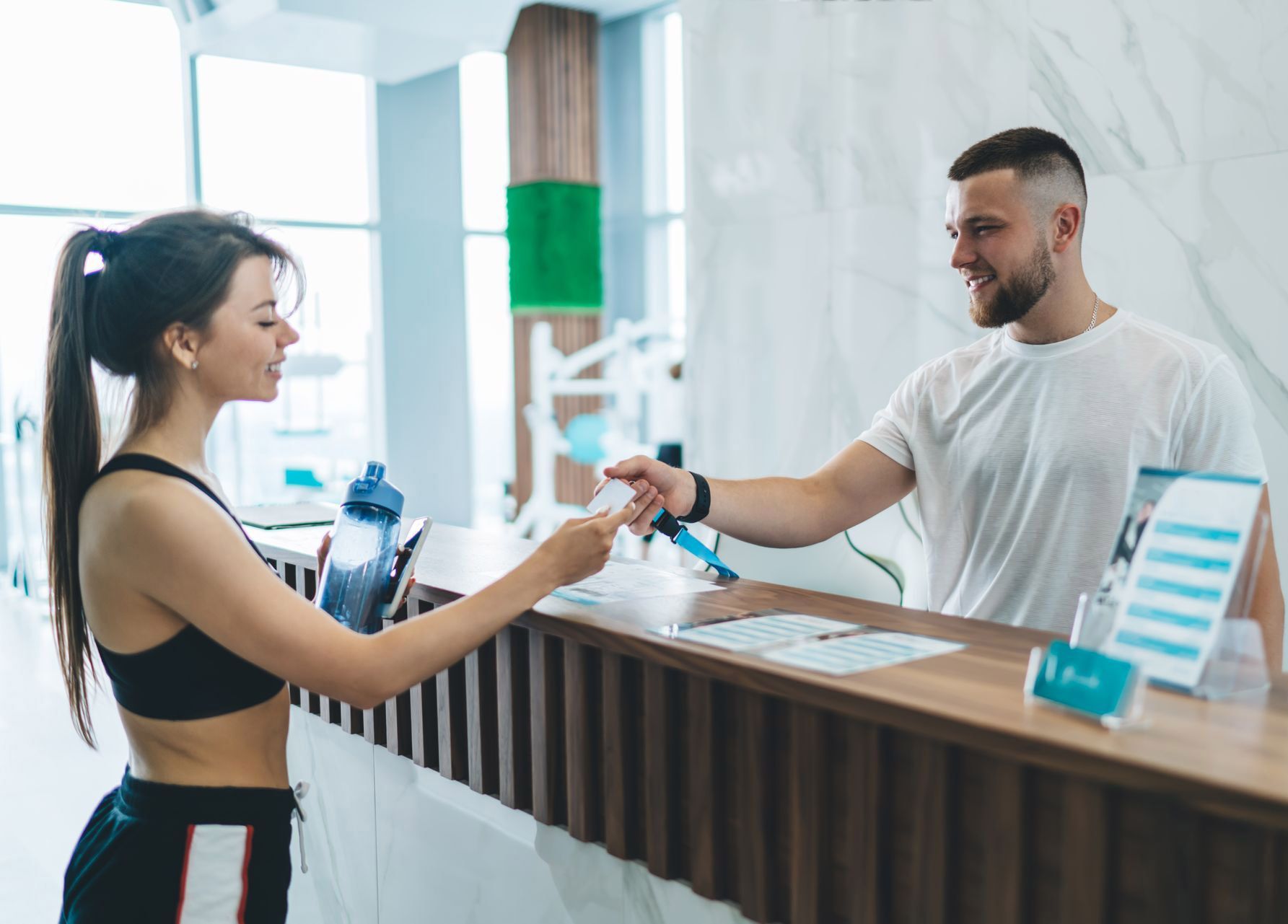 Woman in workout clothes hands card to gym staff at front desk.