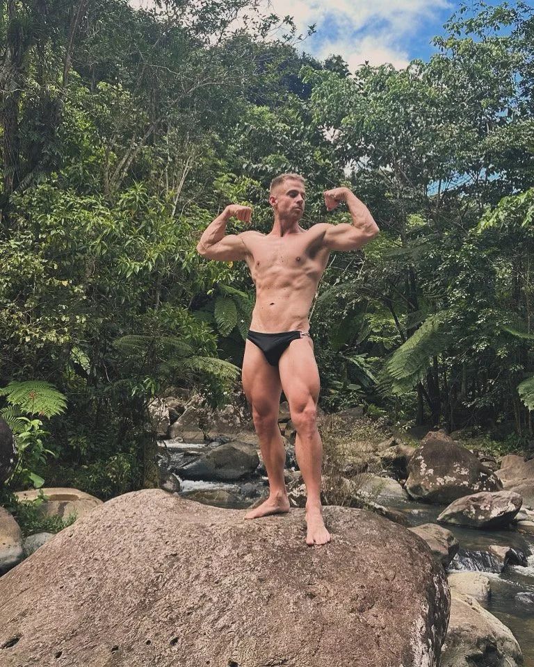 Man flexing muscles in black swimsuit, standing on a rock by a stream, surrounded by lush green foliage.