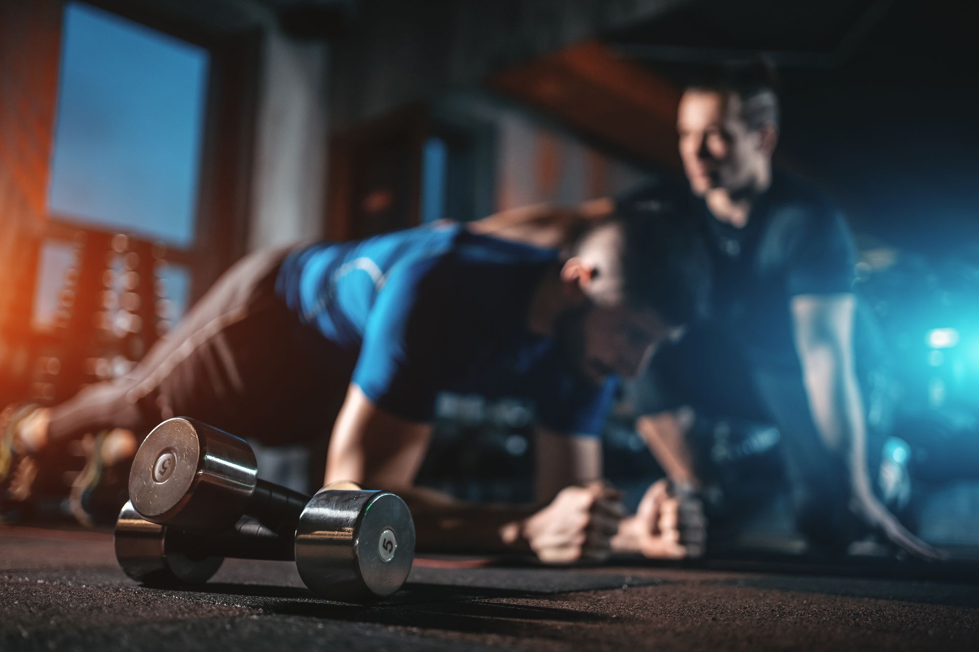 Man doing push-ups with a personal trainer in a gym. Dark setting, weights in foreground.