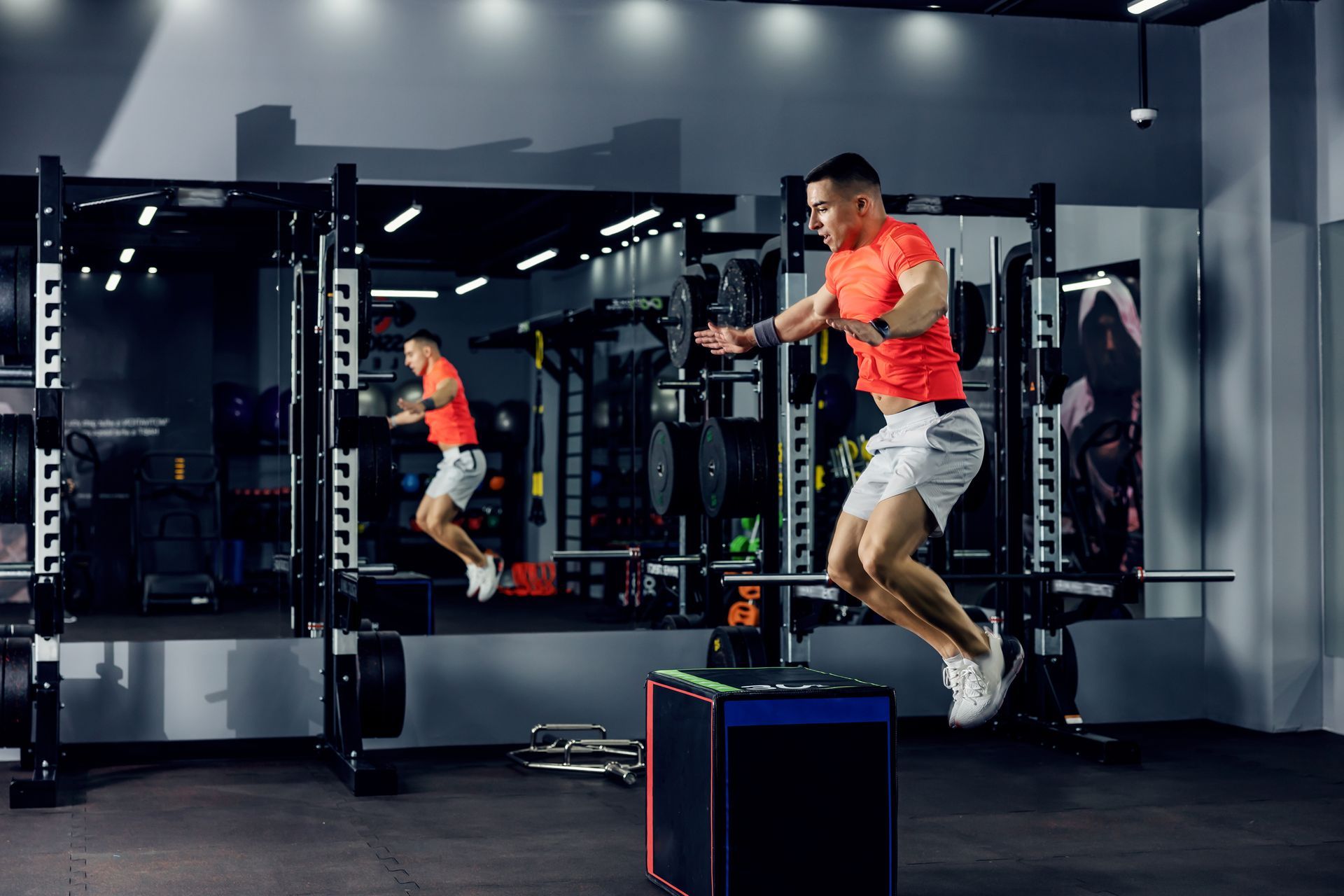 Man in red shirt jumping onto a box in a gym, another person is seen in the mirror.