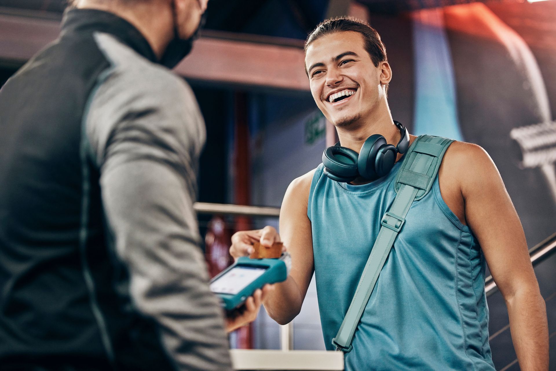 Man paying at gym entrance, smiling, blue tank top, headphones, bag; another person, mask, card reader.