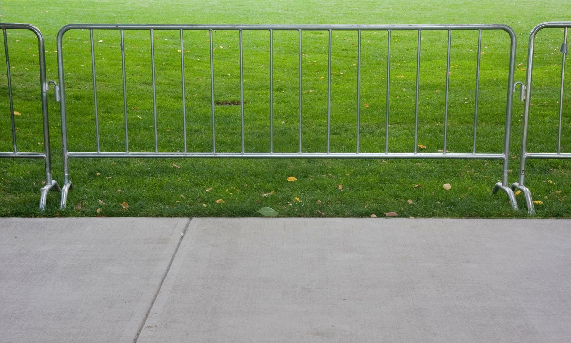 A metal barrier is sitting on the sidewalk in front of a grassy field.