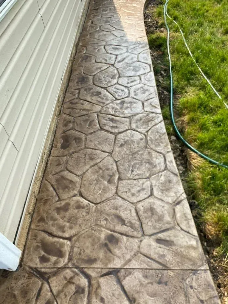 Stamped concrete walkway beside a house, with grass and a green hose along the edge.