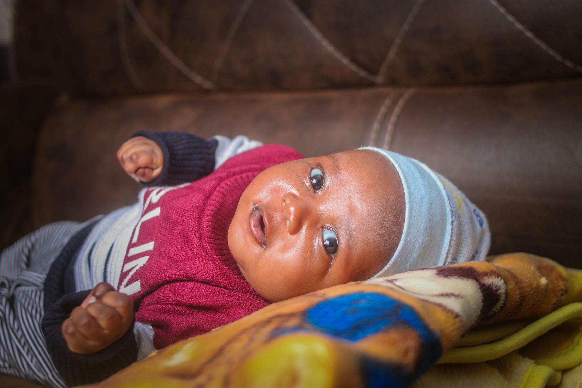 Baby wearing a hat and red sweater, lying on a blanket on a brown couch.