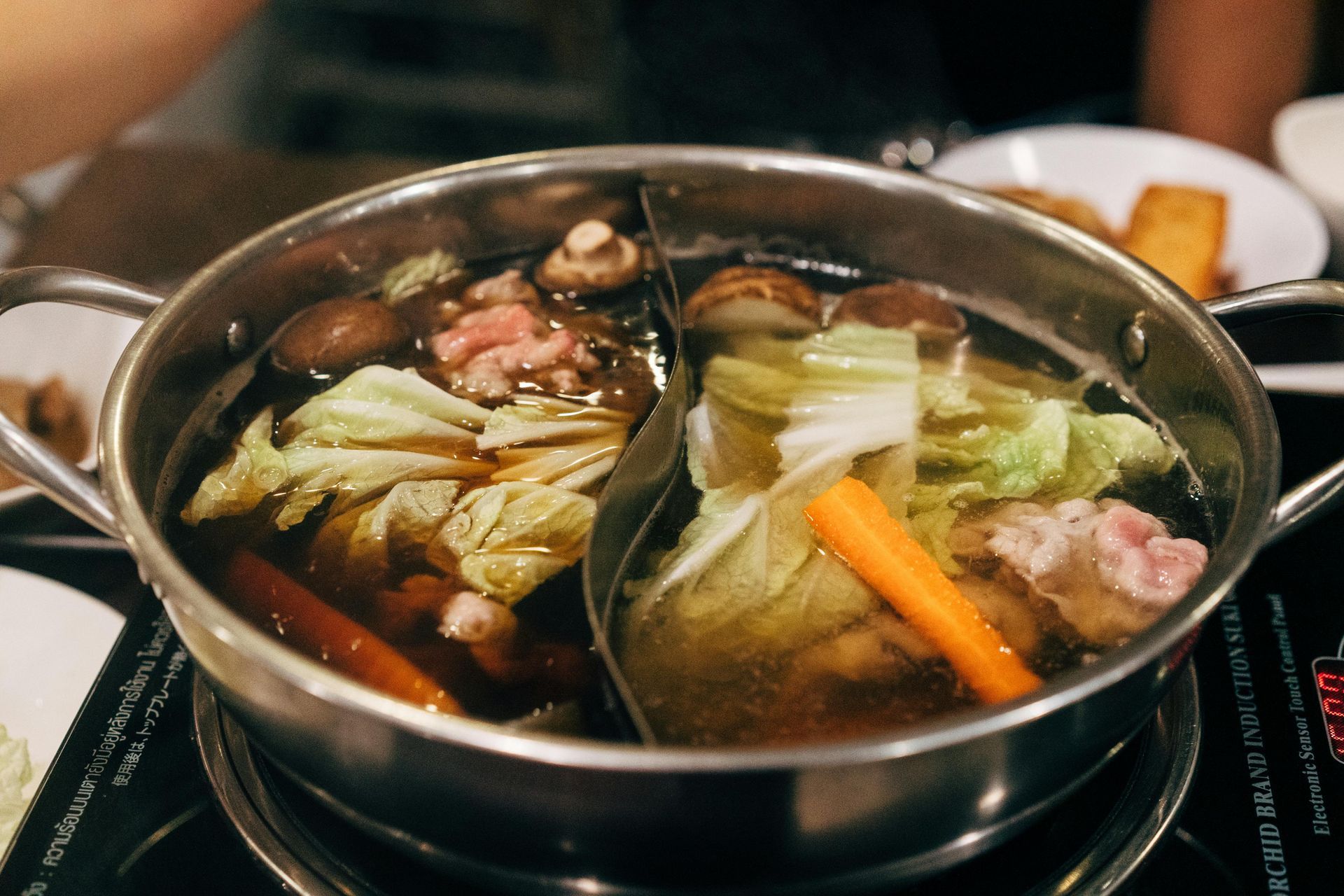 Hot pot with broth, vegetables, and meat, in a divided stainless steel pot, on a burner.