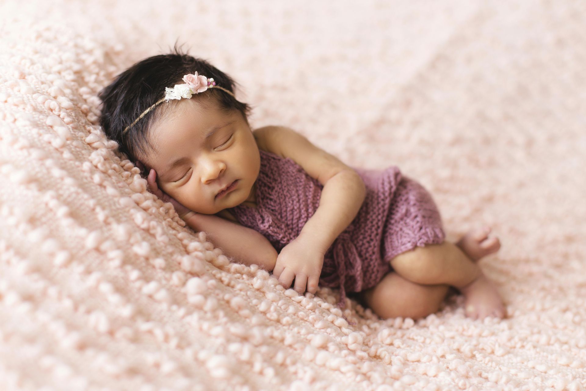 Sleeping baby in a pink knitted outfit and floral headband, lying on a pink textured blanket.