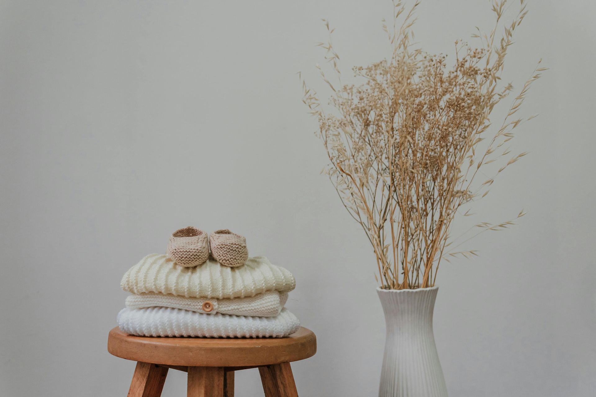 Stack of cream sweaters and baby booties on a wooden stool next to a vase of dried flowers.