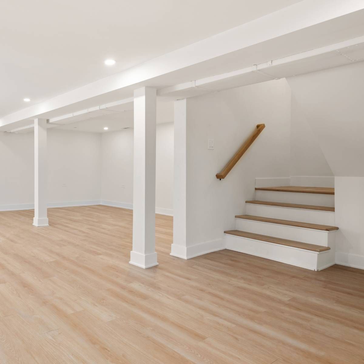 Empty, bright basement with light wood floors, white walls, support beams, and a staircase.