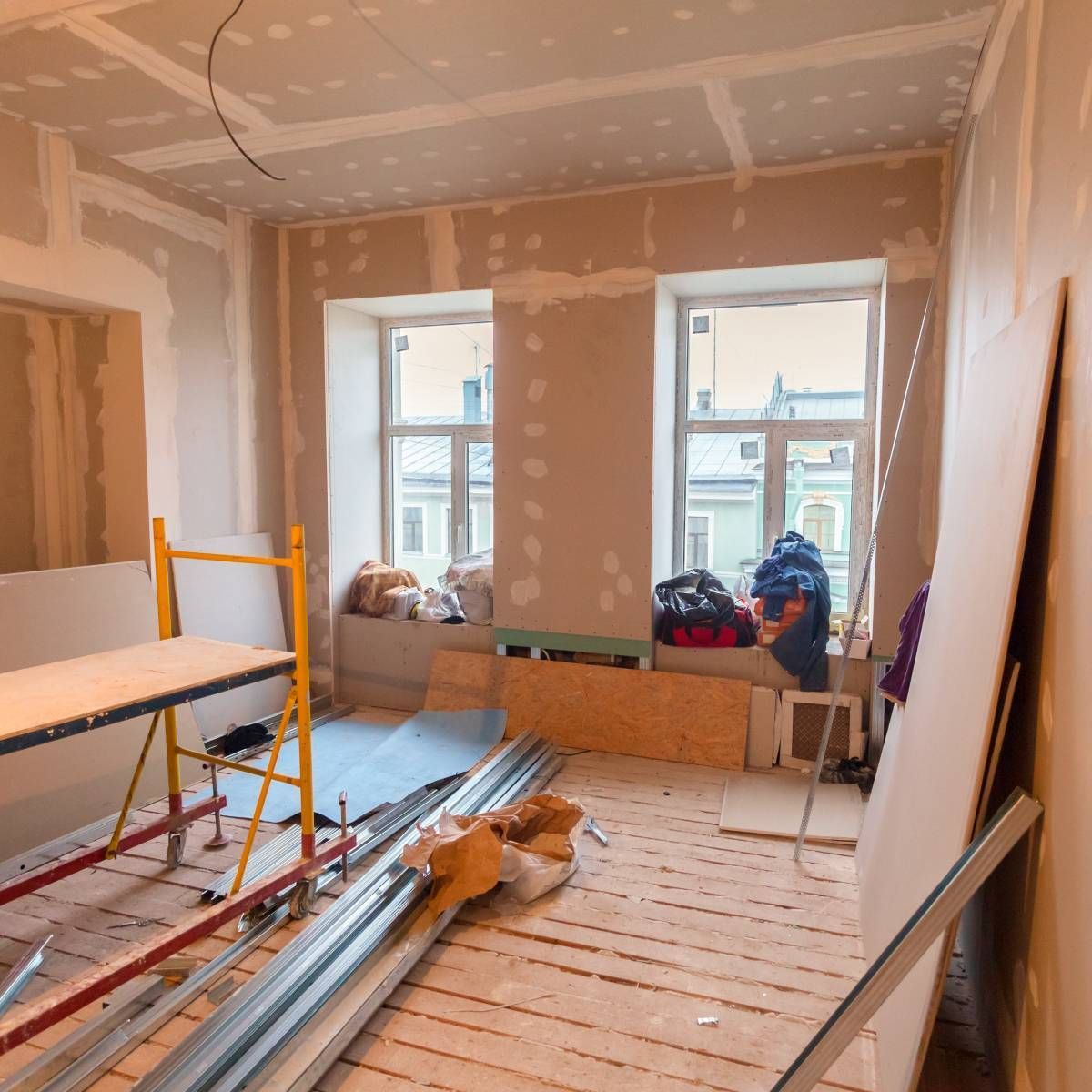 Room under construction with exposed drywall and building materials. Two windows, scaffolding, and wood floor visible.