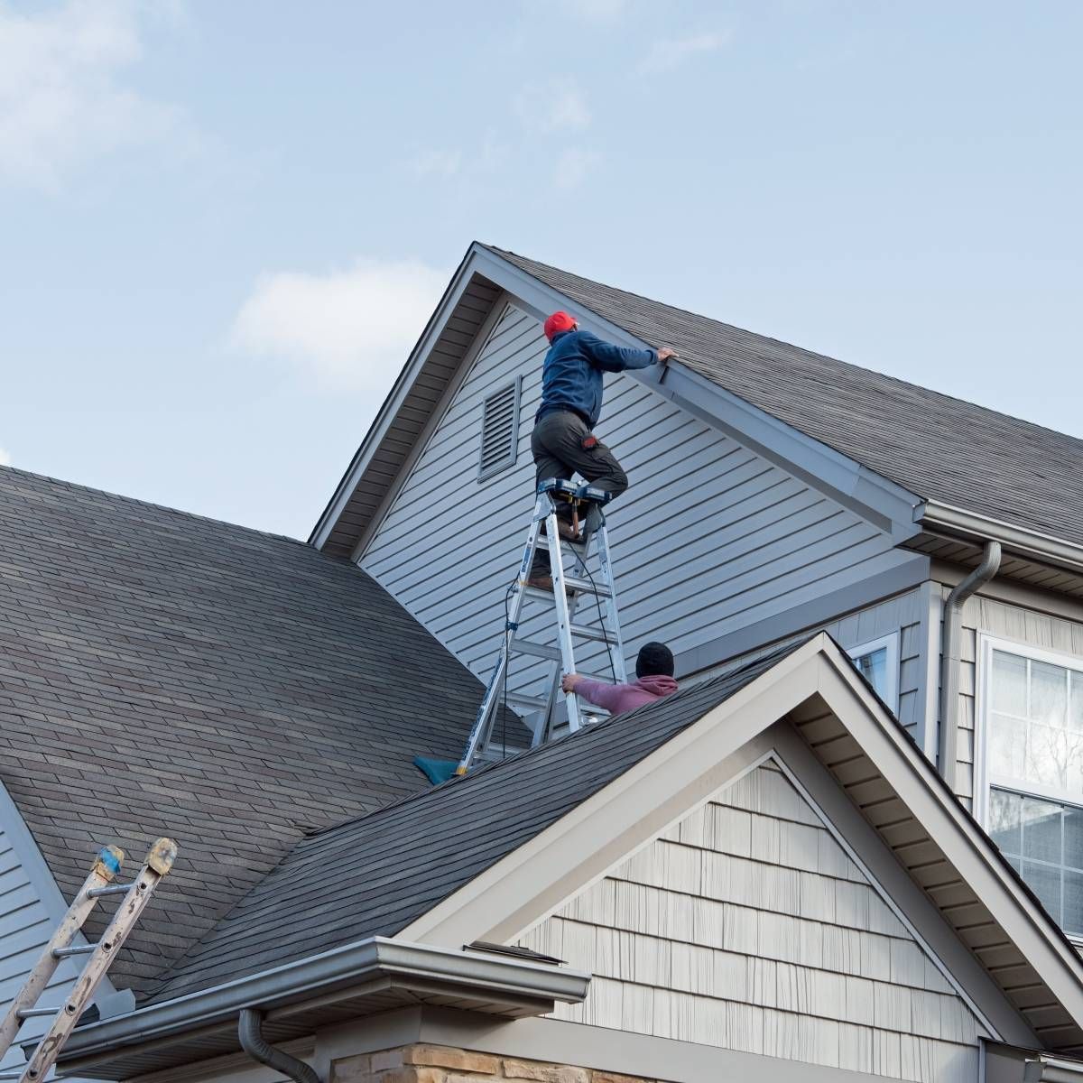 Two people on ladders working on a house roof; one high up, one low; overcast sky.