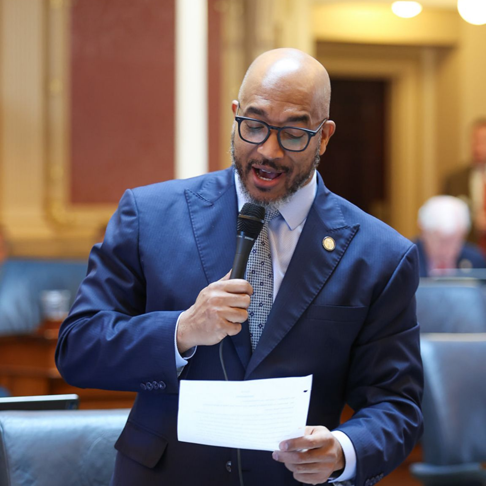 Delegate Mike Jones in a blue suit speaks into a microphone, holding papers in a legislative chamber.