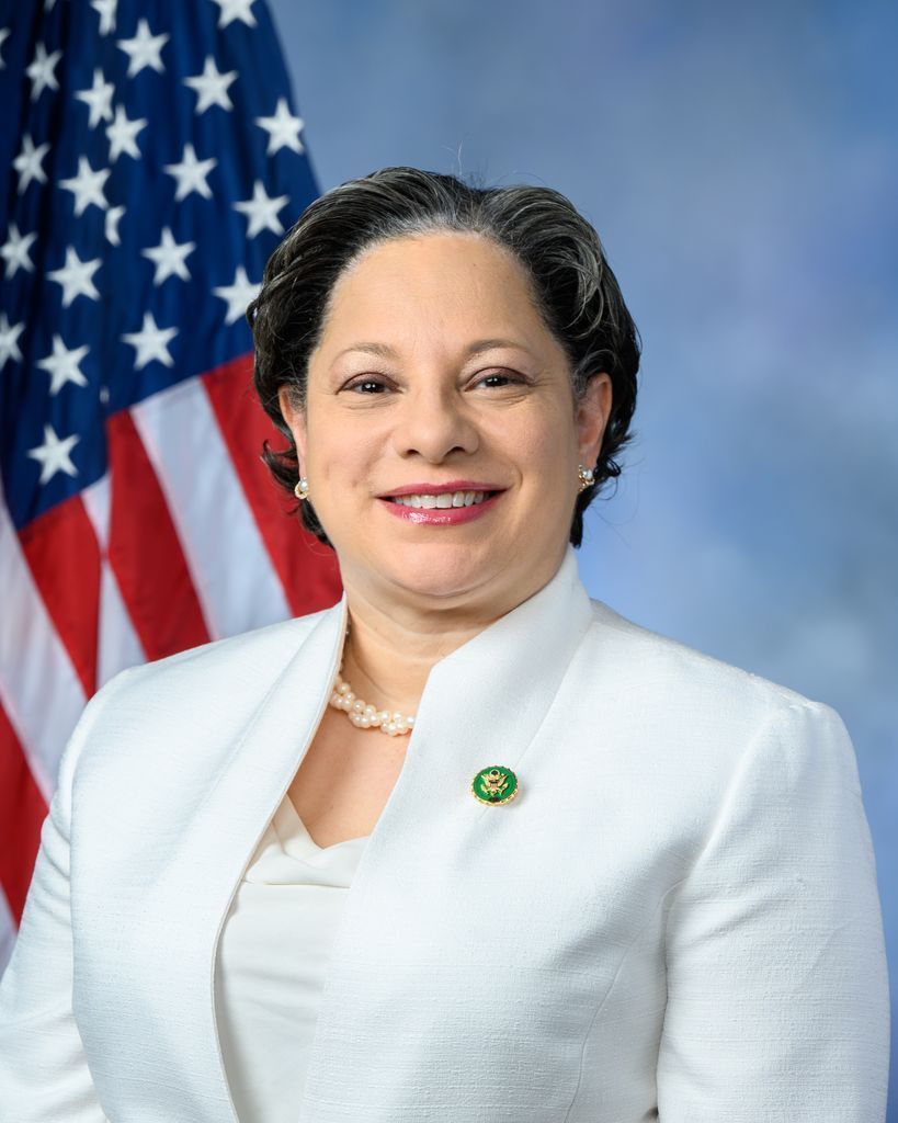 U.S. Representative Jennifer McClellan in a white suit is standing in front of an american flag.