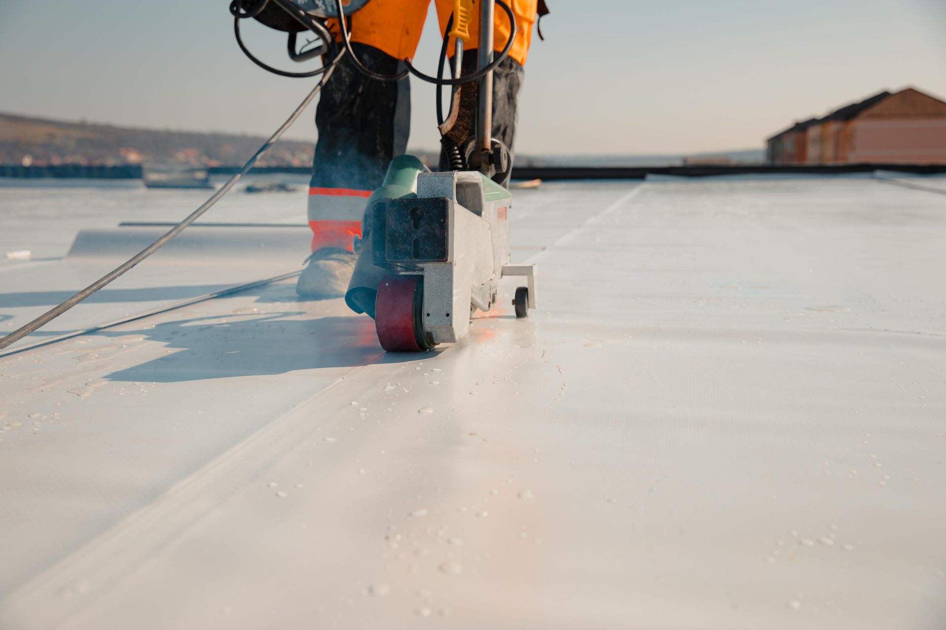 Person in orange workwear welding a white roof seam with a machine, outdoors on a sunny day.