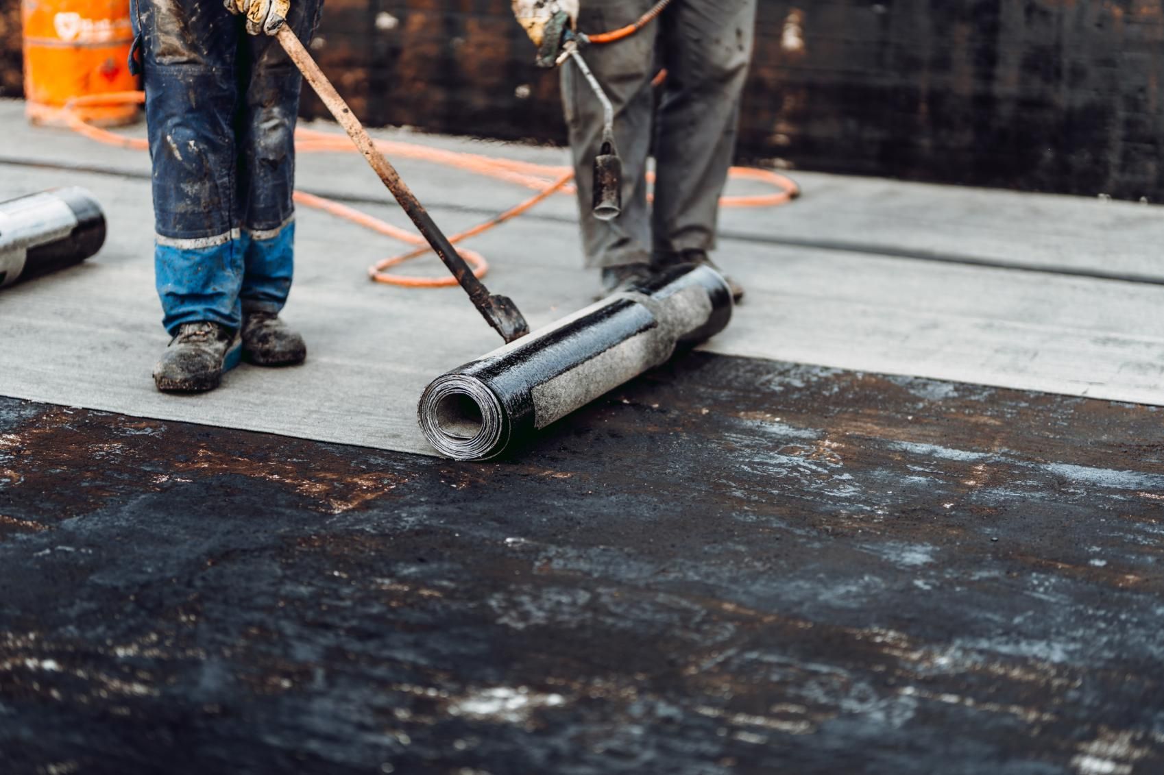 Workers rolling out and smoothing black roofing material on a flat surface, wearing protective gear.