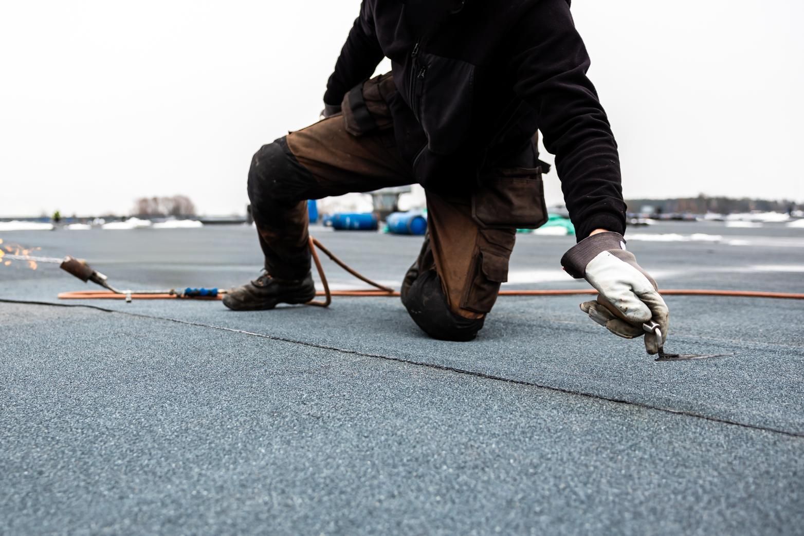 Roofer kneeling on a gray, flat roof, using a torch and tool to seal a seam.