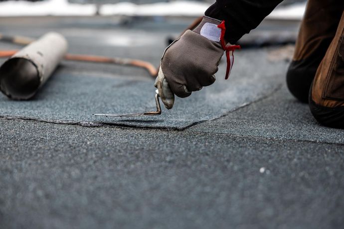 A person wearing gloves uses a tool to work on a flat, dark-colored roof.