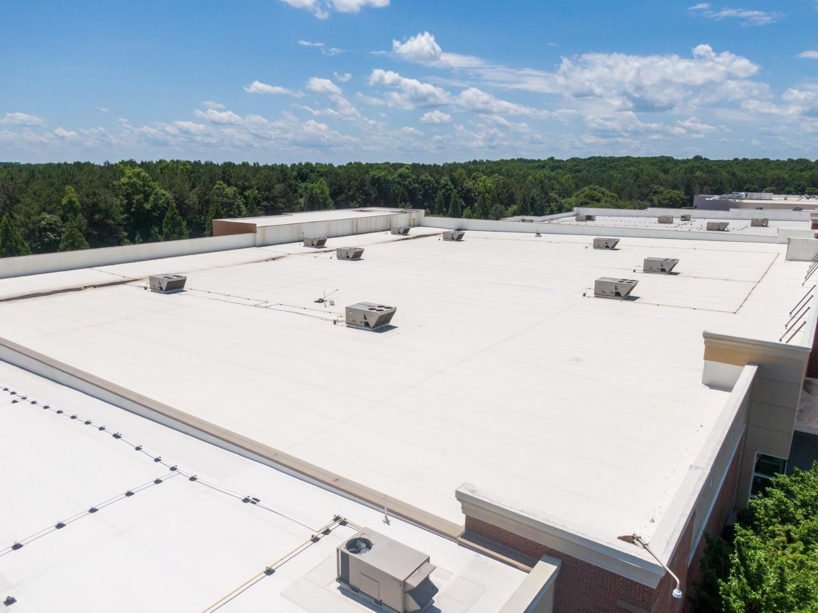 White commercial roof with several HVAC units, blue sky, and treeline in the background.