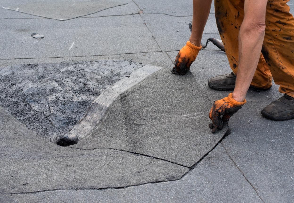 Person in orange overalls repairing a black asphalt roof, placing a patch over a hole.