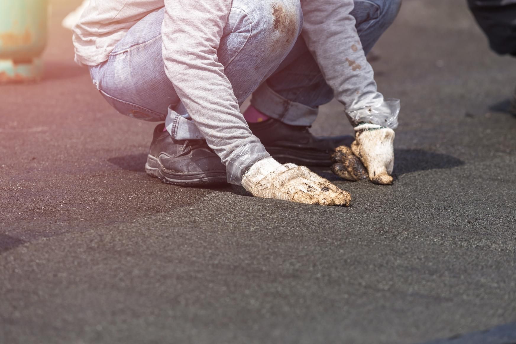 Person in work clothes crouches, touching dark surface with dirty gloved hands.