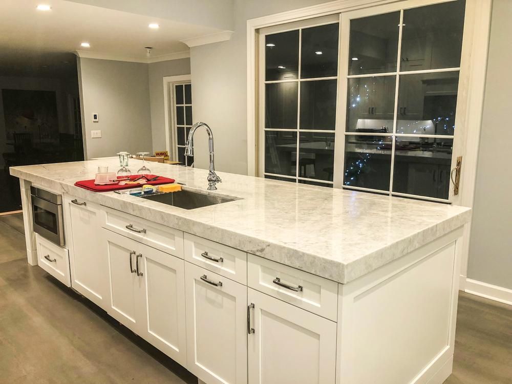 White kitchen island with a marble countertop and a stainless steel sink, facing a large window.