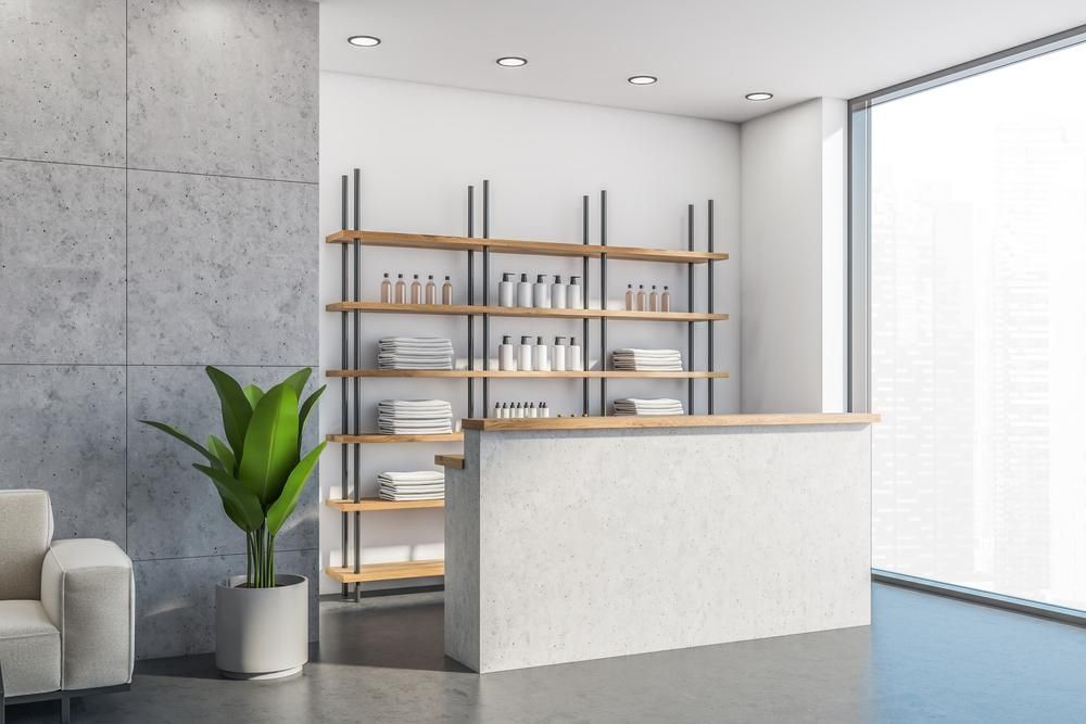A minimalist reception area with a white counter, shelving with products, a large plant, and a concrete wall.