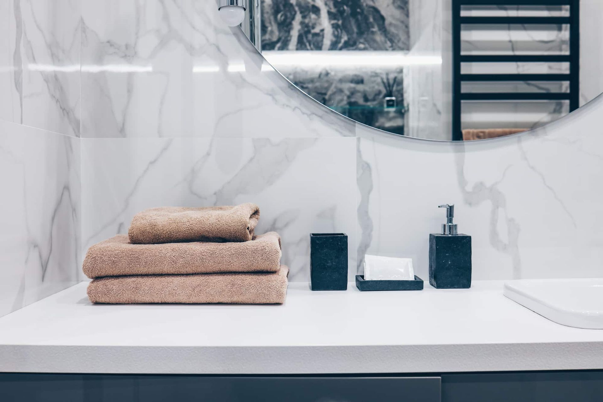 Bathroom vanity with tan towels, soap dispenser, and accessories against marble-look tile.