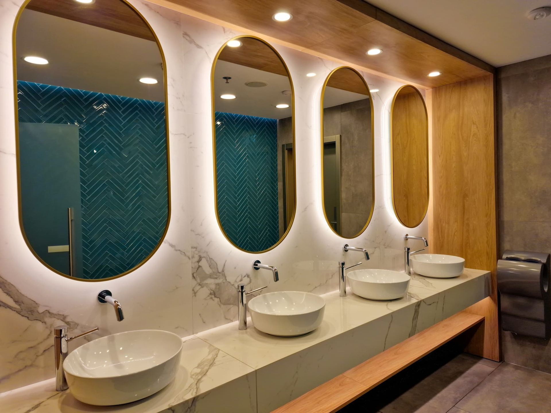 Modern public restroom with oval mirrors, white sinks, and teal tiled walls.