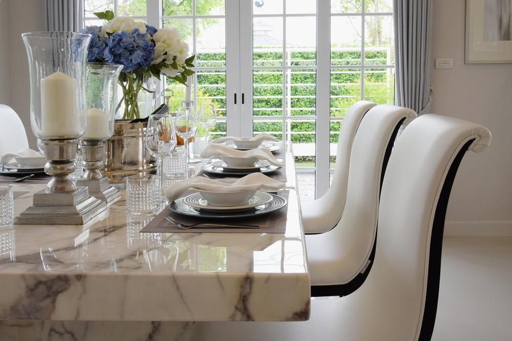 Dining room table set for a meal. Marble top, white chairs, and large window in the background.