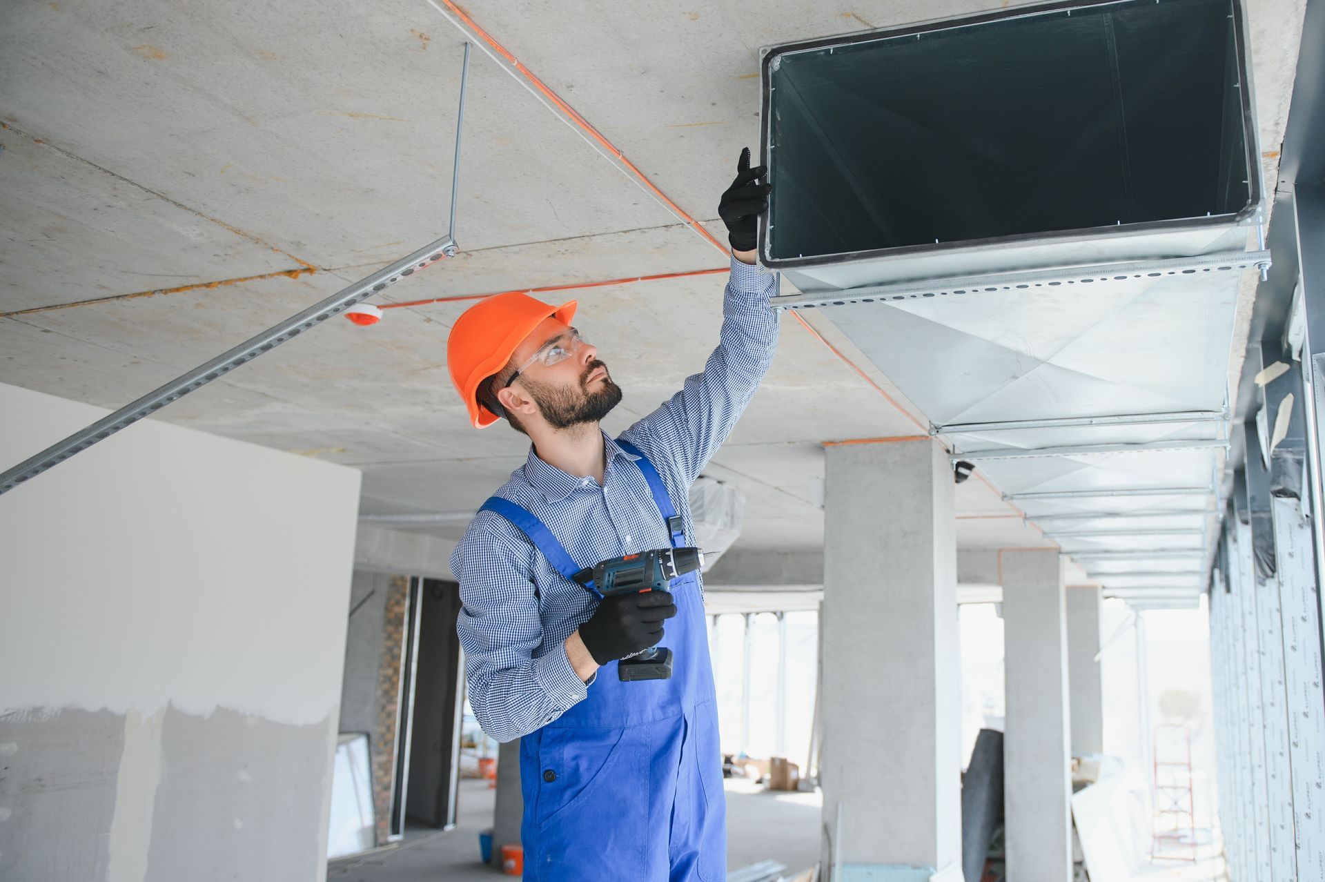 Construction worker in blue overalls and orange helmet installing ductwork, holding a drill. Construction worker in blue overalls and orange helmet installing ductwork, holding a drill.