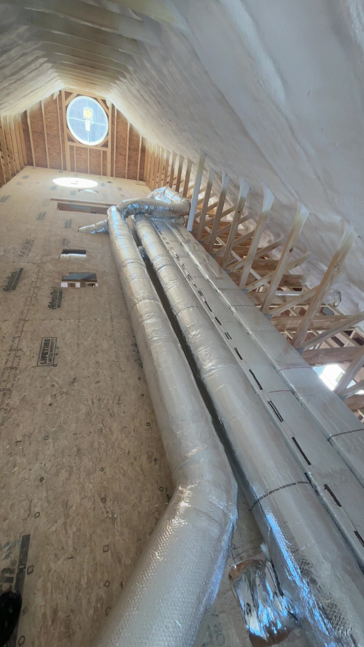 Attic with insulated HVAC ductwork running lengthwise. Wooden framing visible. Circular window at far end.