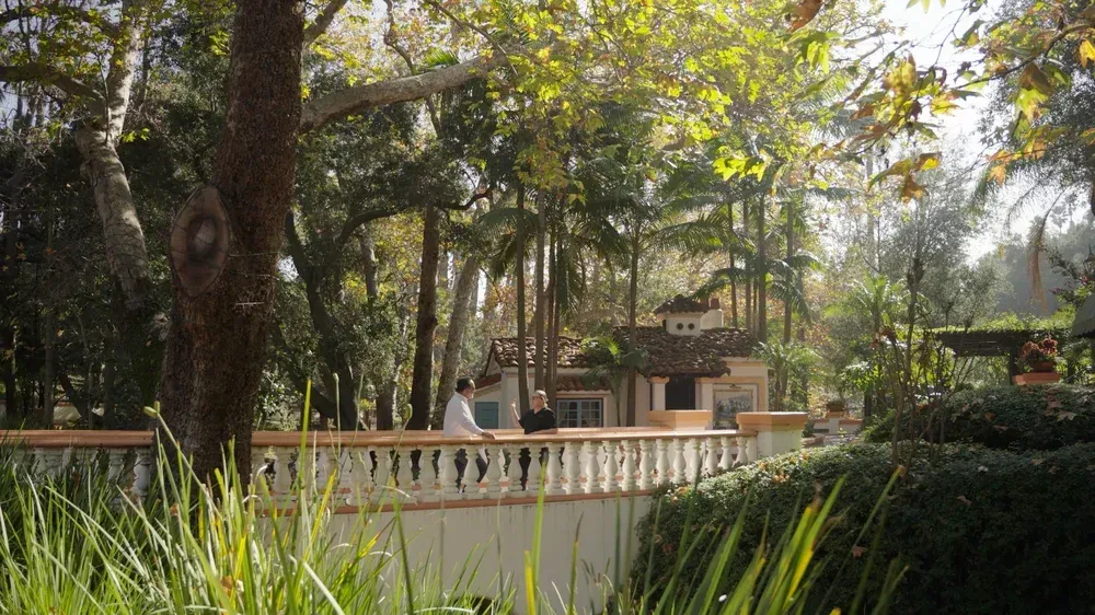 A bridge with two people overlooking a Spanish-style house surrounded by trees and greenery.