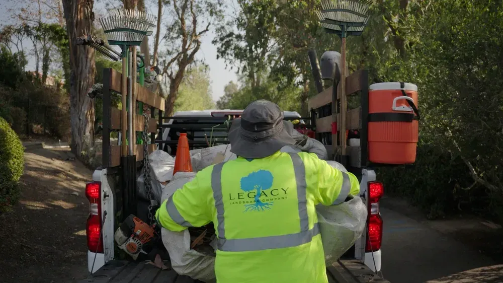 A person in a reflective vest loads a truck bed with landscaping supplies, trees in the background.