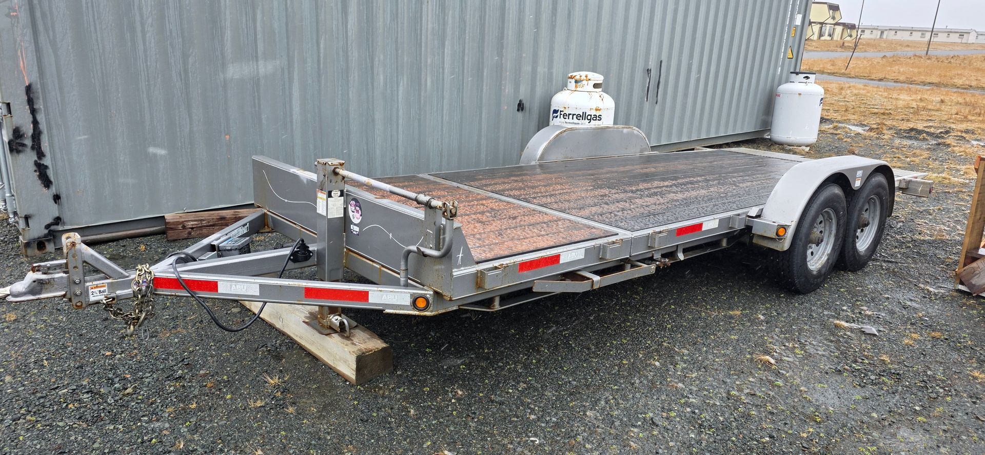 14,000lb tilt deck equipment trailer A gray car trailer parked on gravel, with a metal building and propane tanks in the background.
