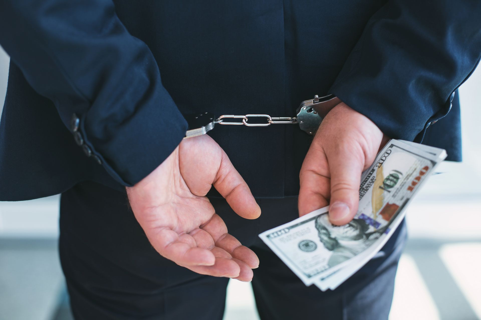 Man in suit, handcuffed, holding a wad of US currency behind his back.