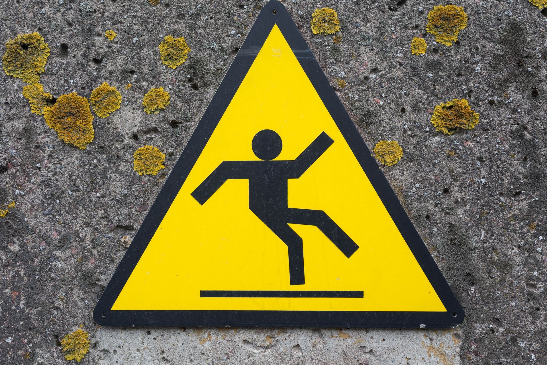 Yellow triangular warning sign depicting a person slipping; set against a concrete wall.