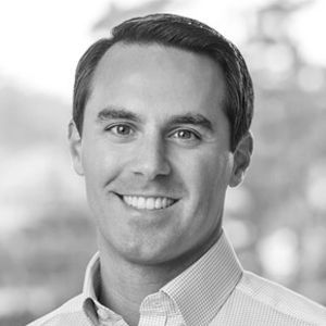 Man in a light-colored shirt, smiling directly at the camera. Black and white, studio shot.