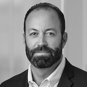 Man with dark hair and beard, wearing a suit jacket and open-collared shirt, looking at the viewer.