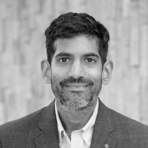 Man with a beard wearing a blazer, smiling in front of a textured wall.
