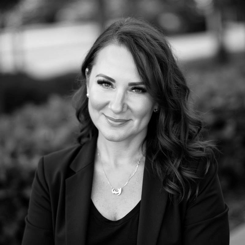 Woman in a black blazer smiles, posing outdoors with blurred greenery background.