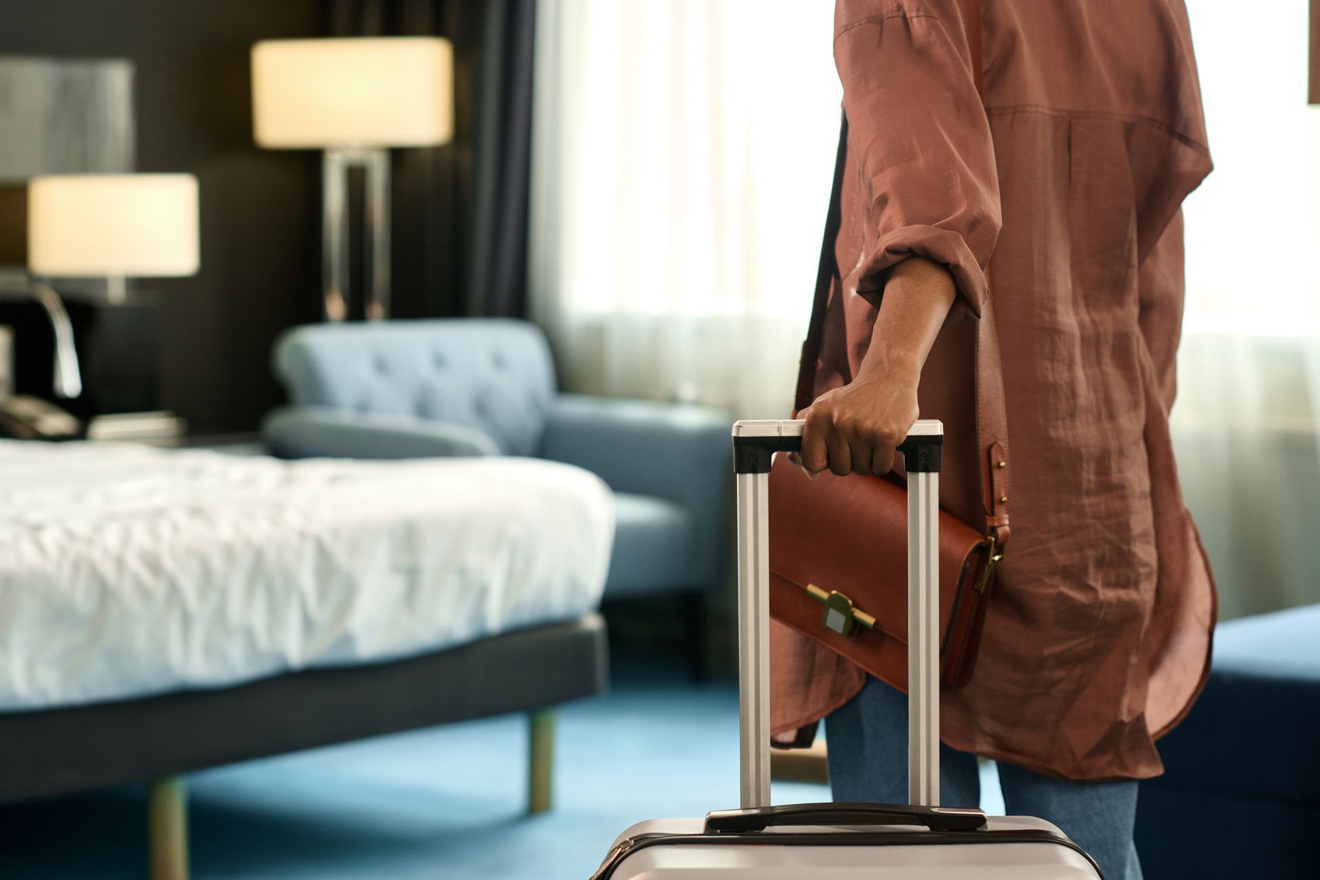 Person entering hotel room, holding suitcase. Room has bed, chair, and lamps.