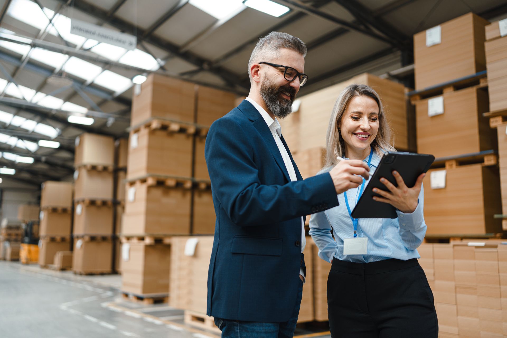 Man in blue blazer points at tablet held by woman in warehouse.