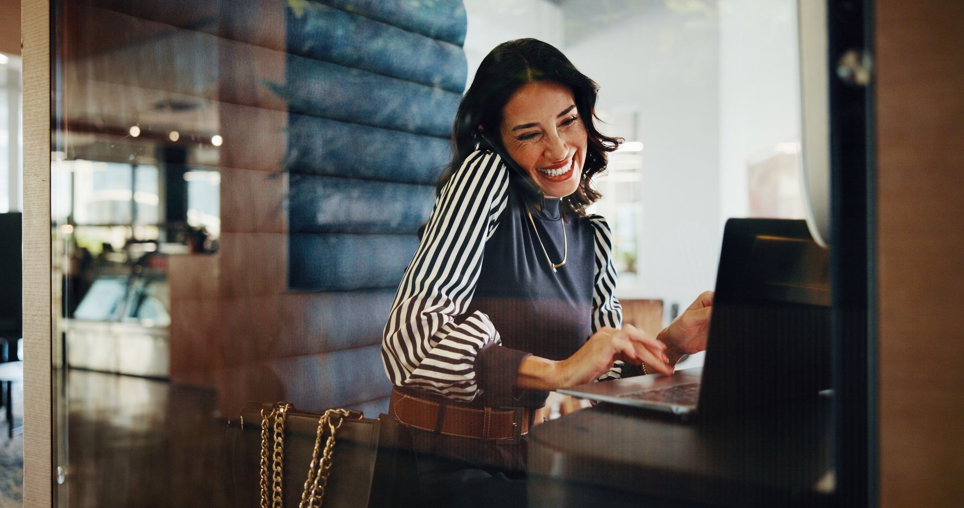 Woman on phone smiles, using laptop at a cafe. She wears a striped shirt.