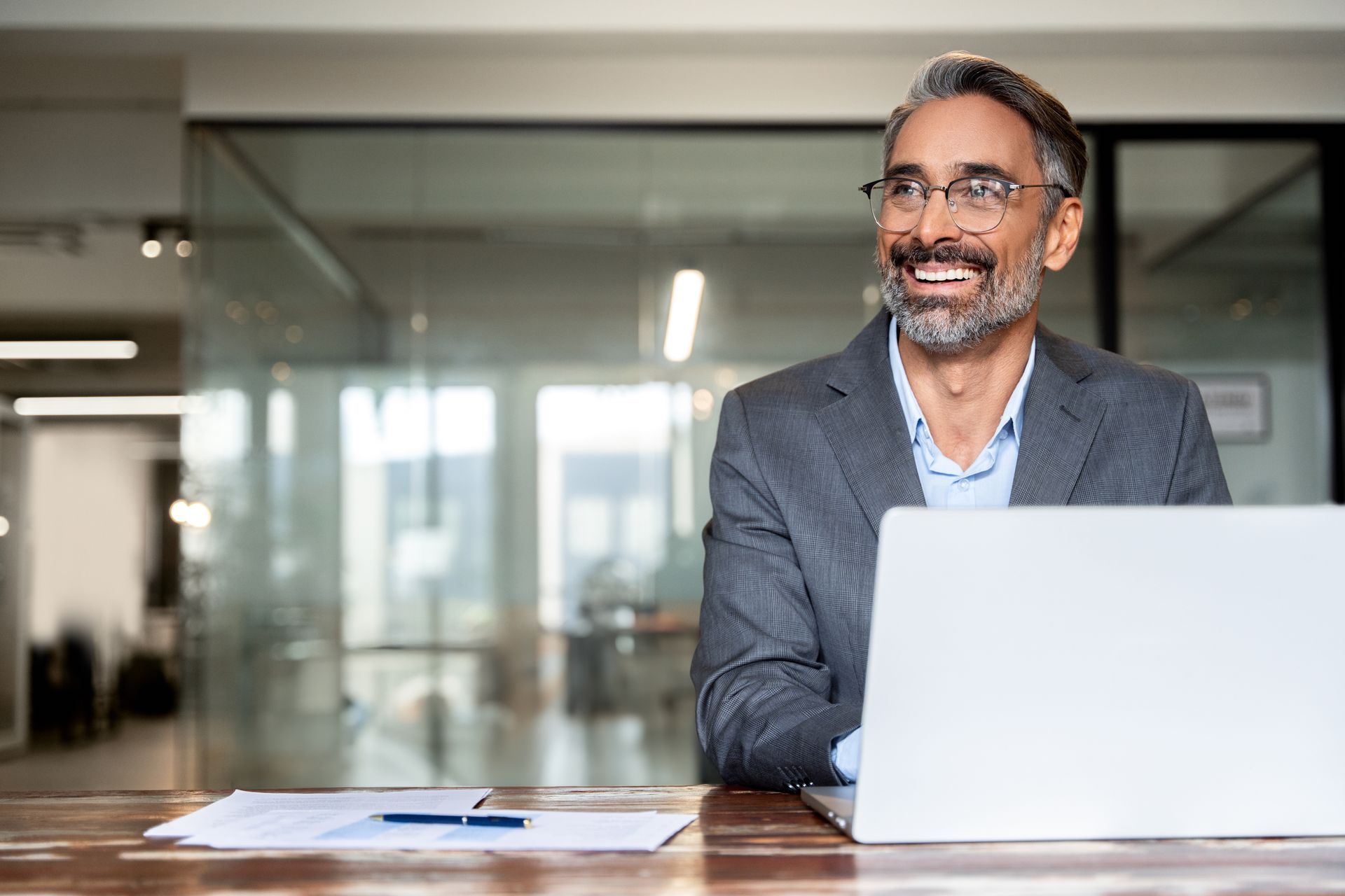Smiling man in a suit at a desk with a laptop and papers, in an office setting.