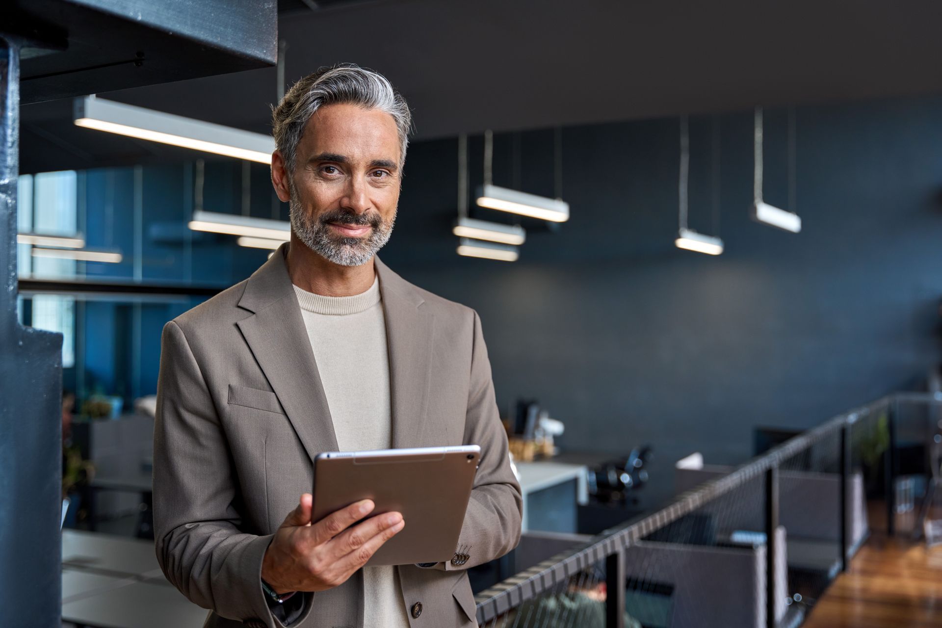 Man in tan blazer holding tablet, smiles in modern office setting.