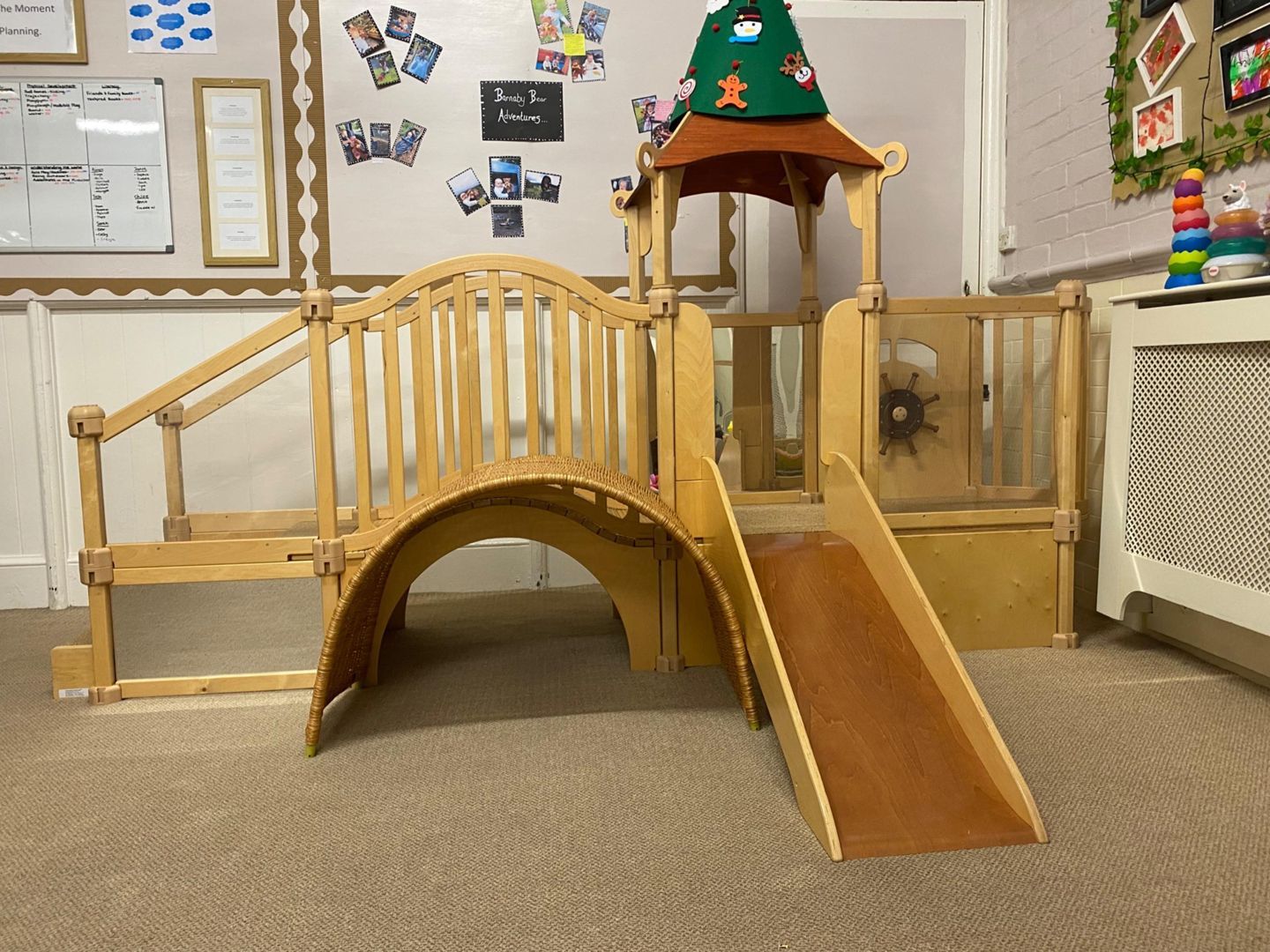 Wooden play structure with a slide, steps, and a bridge in a room.