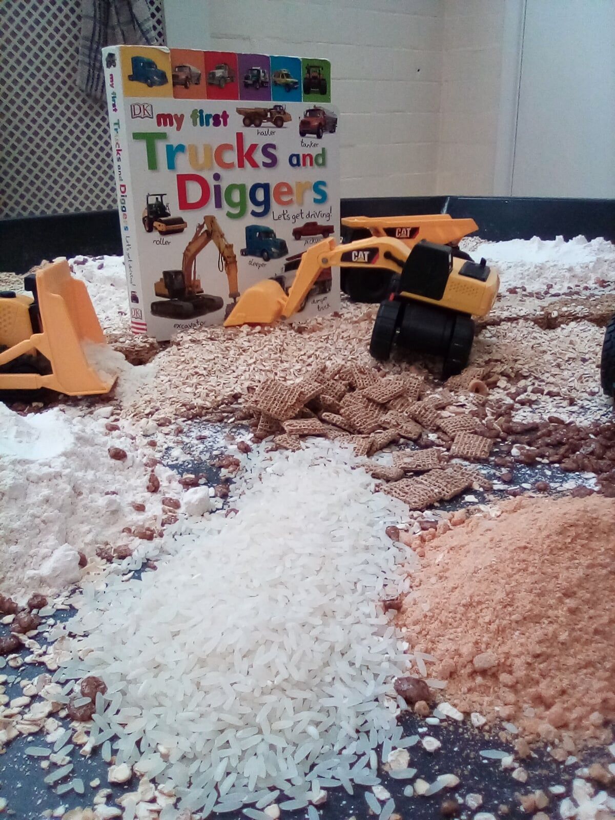 Construction-themed sensory bin. Trucks and diggers surrounded by rice, flour, and sand. Book titled