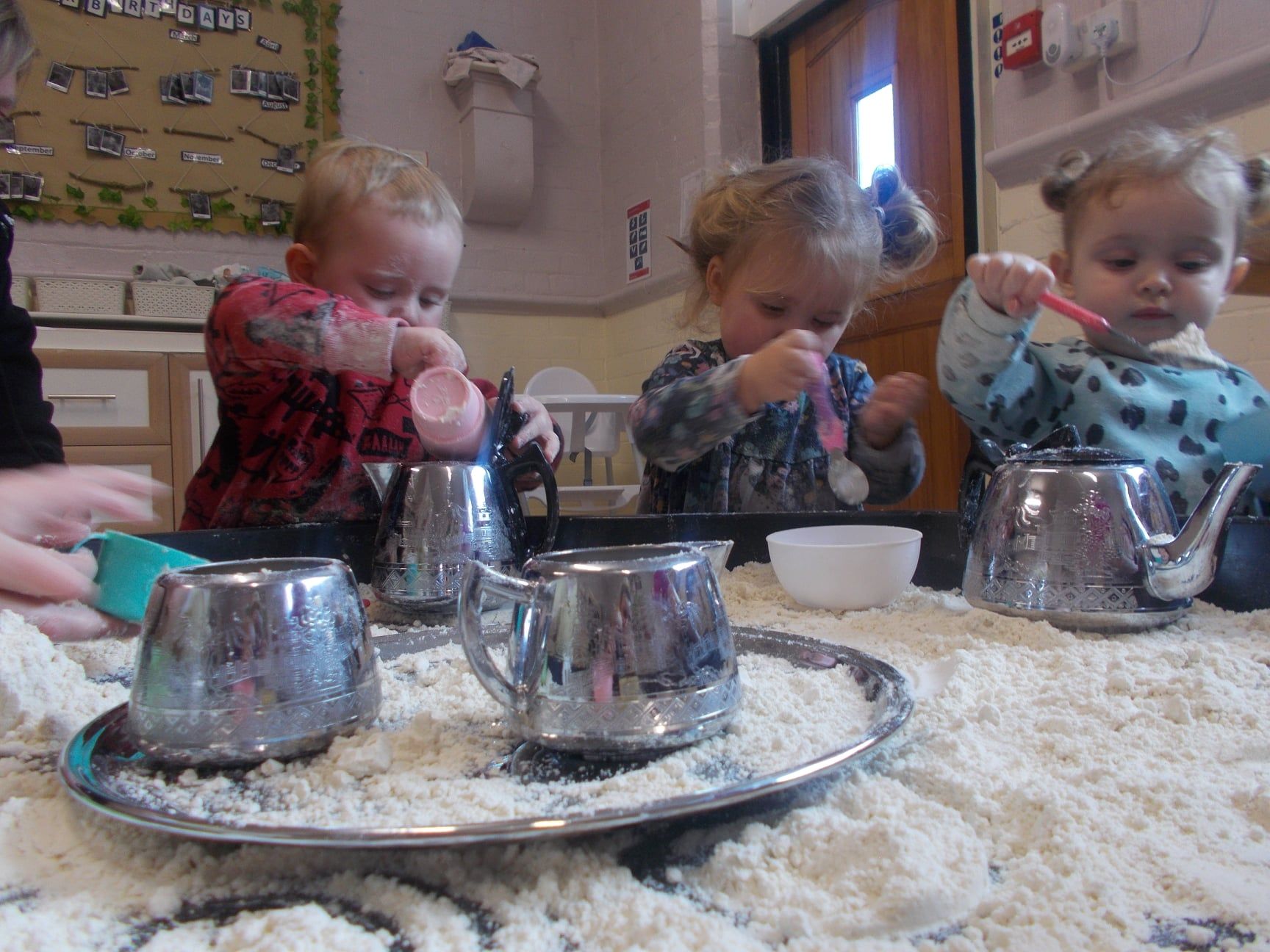 Children playing with flour, silver teapots, and spoons on a table.