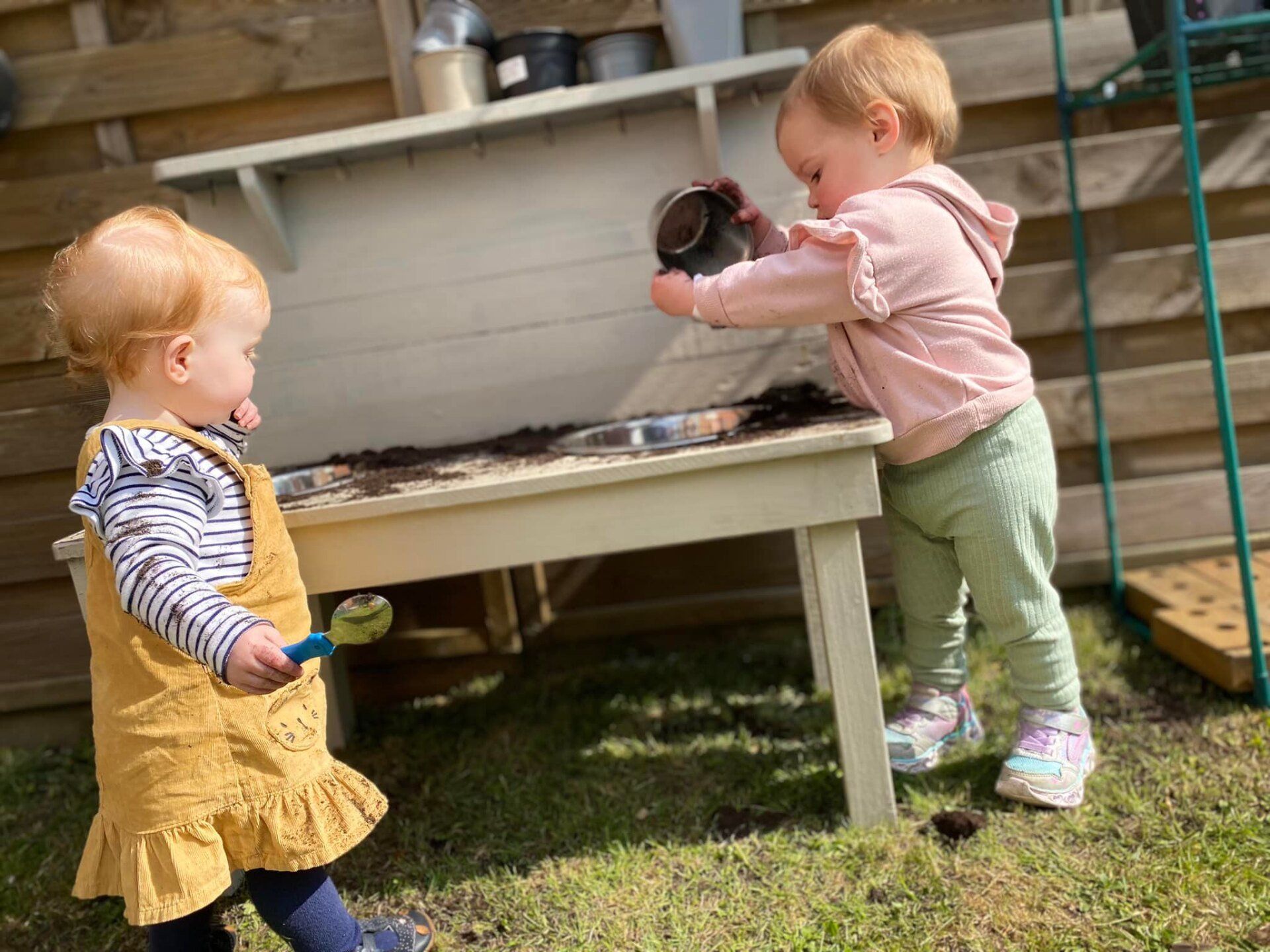 Two toddlers playing with dirt in a sandbox. One pouring, other holding a toy spoon. Outdoors.