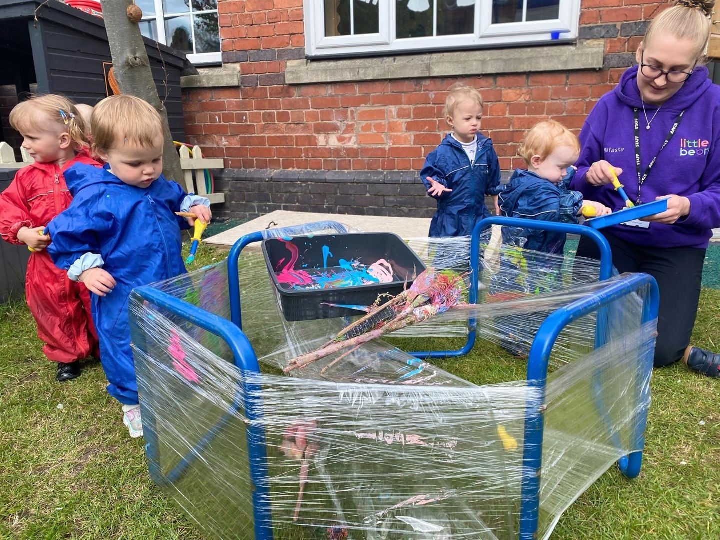 Children in raincoats painting on a plastic-wrapped frame outdoors, supervised by a caregiver.